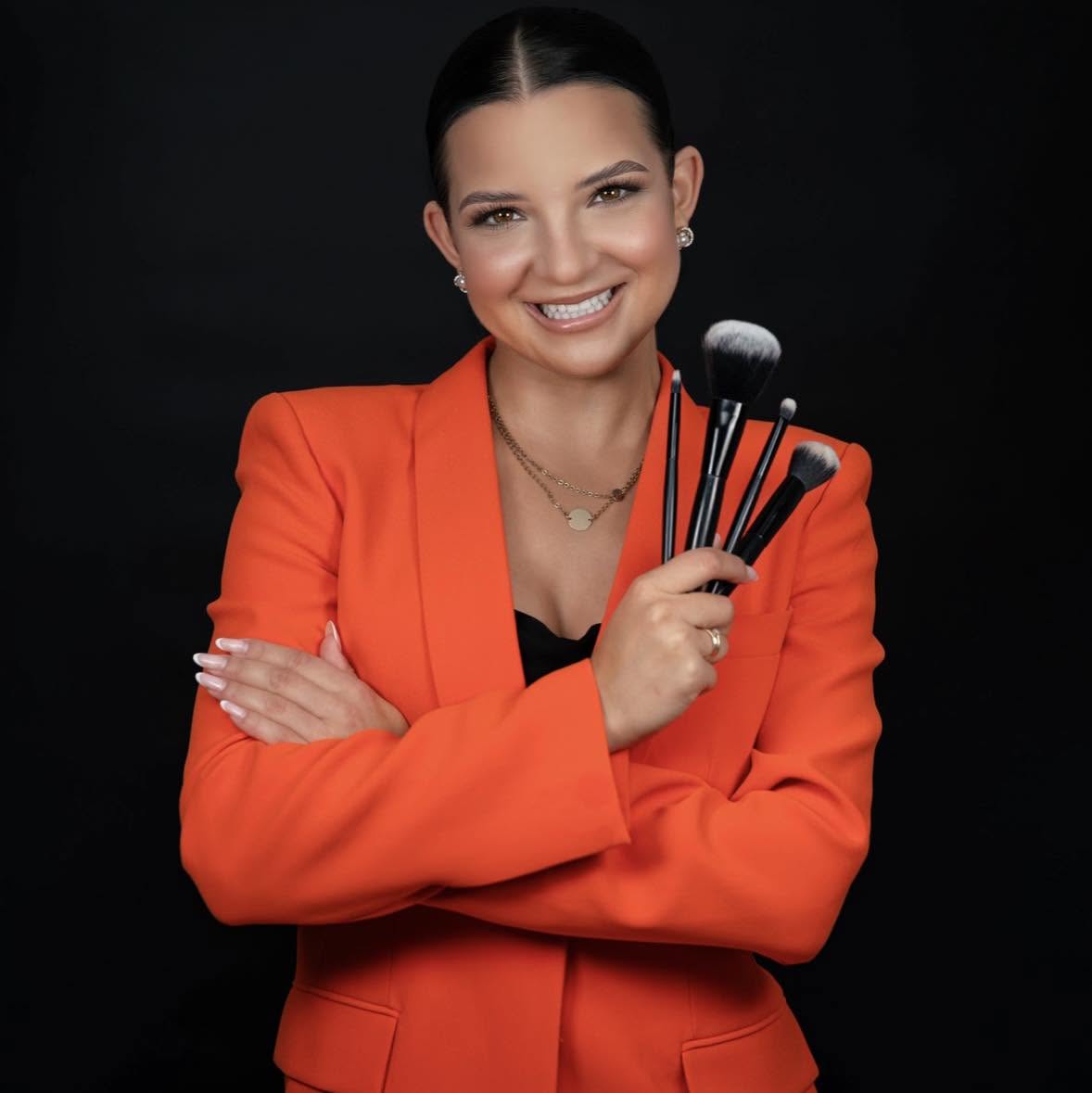 Magda Adamowicz w pomarańczowym żakiecie, uśmiechnięta, trzyma pędzle do makijażu